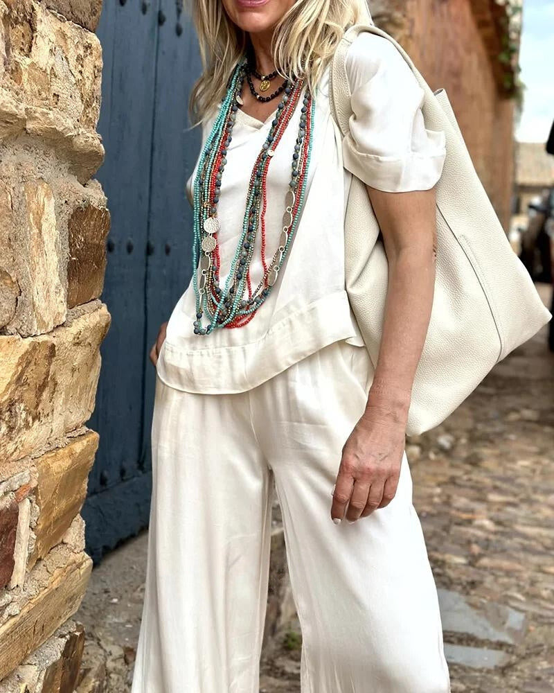 Elegante Frau in weißem Outfit mit langen, bunten Halsketten, posiert vor rustikaler Steinmauer. Modetrend, Sommermode, Boho-Chic.
