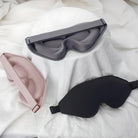 Drei Schlafmasken aus Stoff in Schwarz, Grau und Rosa auf weißem Hintergrund.