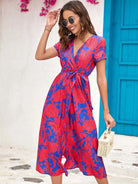 Frau in rotem, geblümtem Sommerkleid vor blauer Tür, trägt Sonnenbrille und Strohtasche. Perfekt für Strandmode und Sommeroutfits.