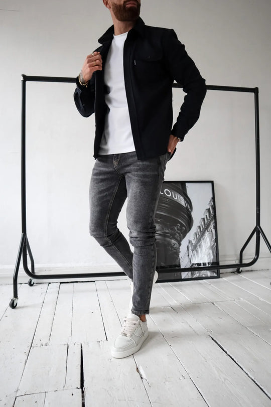 Mann in schwarzer Jacke, weißem T-Shirt und grauen Jeans posiert vor modernem Hintergrund. Trendige Herrenmode, lässiger Stil, Streetwear.