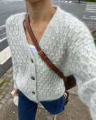 Damen-Strickjacke aus weichem, cremefarbenem Garn mit Knopfleiste und V-Ausschnitt, kombiniert mit Jeans und brauner Umhängetasche.