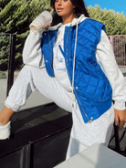 Frau in blauer, ärmelloser Steppweste mit Reißverschluss, kombiniert mit grauem Hoodie und Jogginghose. Modetrend, lässiger Streetstyle, Damenmode.