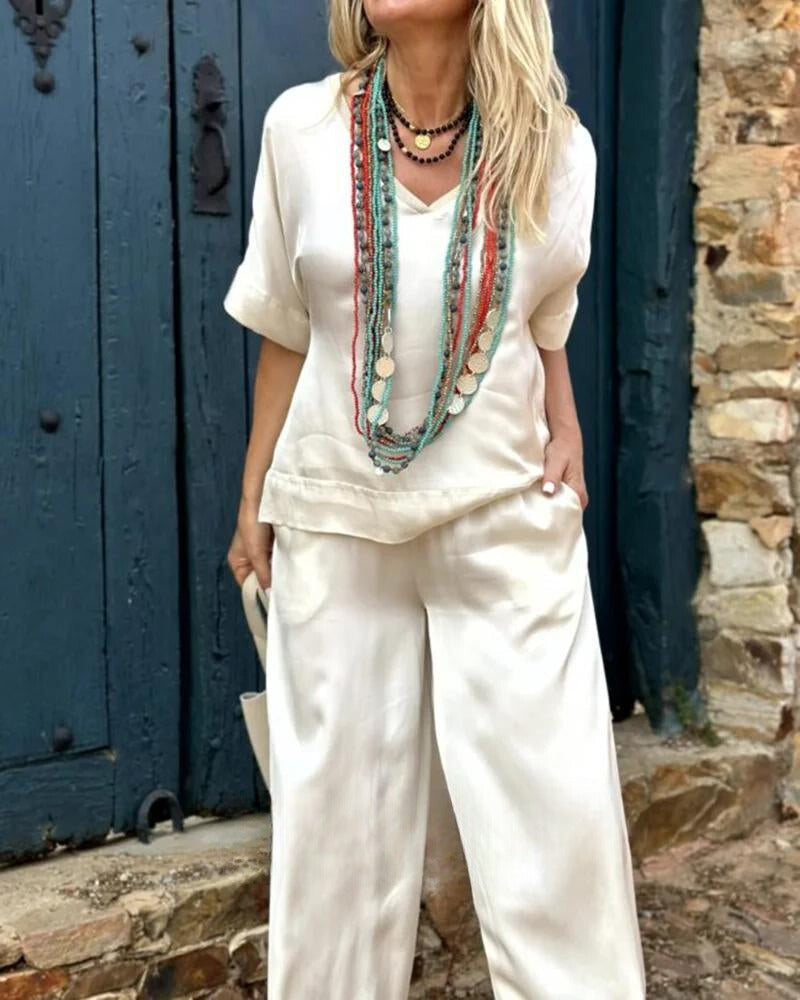 Elegantes Damen-Outfit in Weiß mit lockerer Bluse und Hose, ergänzt durch bunte, lange Halsketten. Perfekt für Sommermode und Boho-Stil.