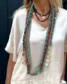 Frau mit weißem Oberteil trägt lange, bunte Halsketten mit Perlen und Münzanhängern. Boho-Schmuck, modische Accessoires, Statement-Ketten.
