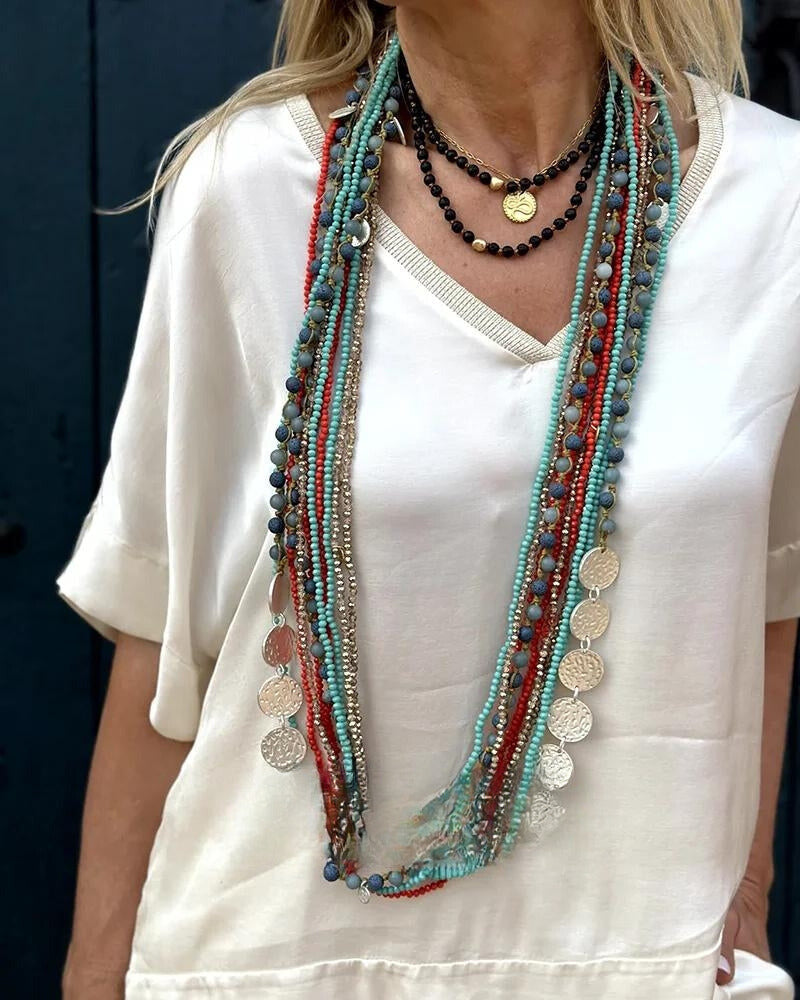 Frau mit weißem Oberteil trägt lange, bunte Halsketten mit Perlen und Münzanhängern. Boho-Schmuck, modische Accessoires, Statement-Ketten.