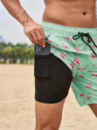Mann in grüner Flamingo-Badeshorts mit Smartphone-Tasche am Strand.
