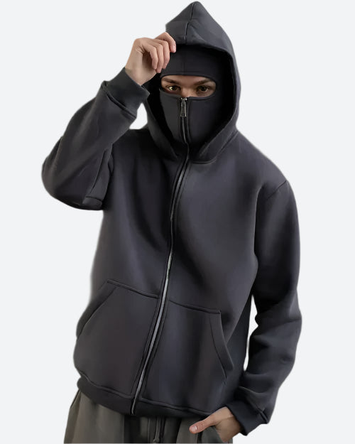 Mann in dunkelgrauem Kapuzenpullover mit Reißverschluss und Maske, urbaner Streetwear-Look.