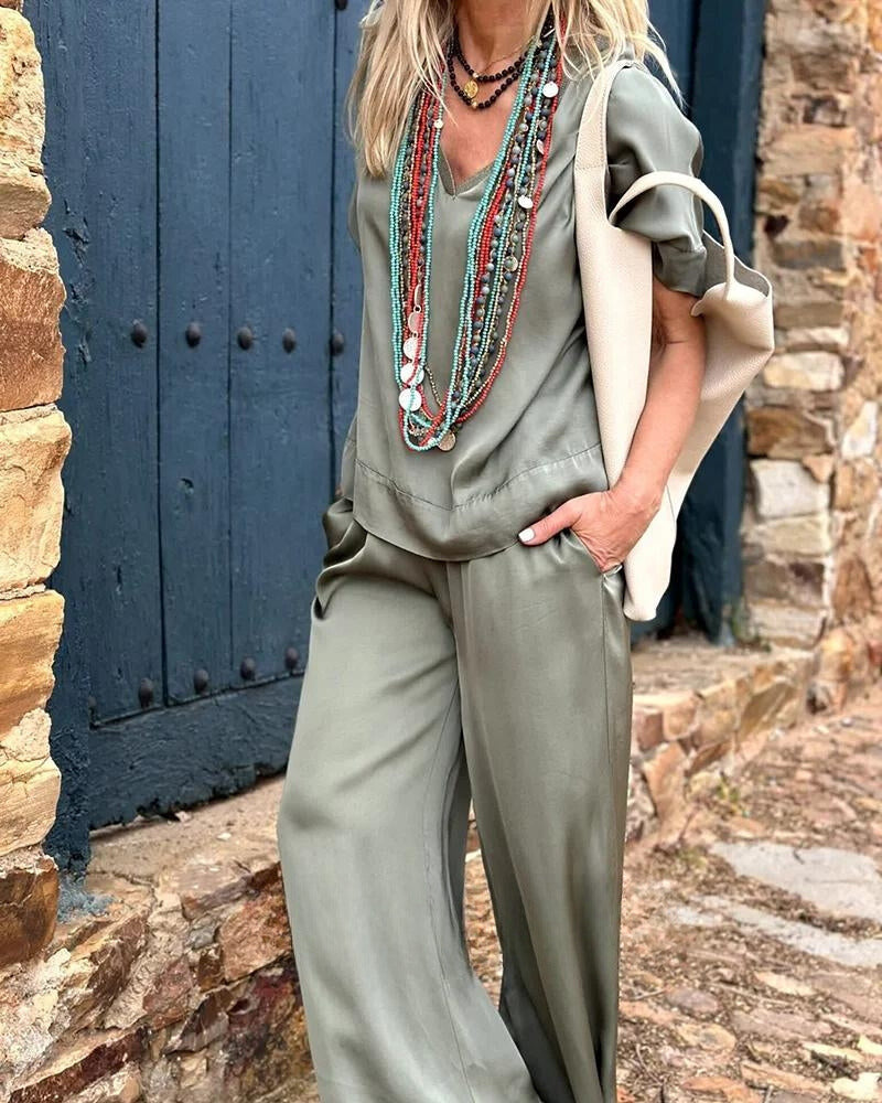 Frau in olivgrünem Jumpsuit mit langen Halsketten, vor rustikaler Steinmauer. Modetrend, lässiger Stil, Boho-Chic, Damenmode.