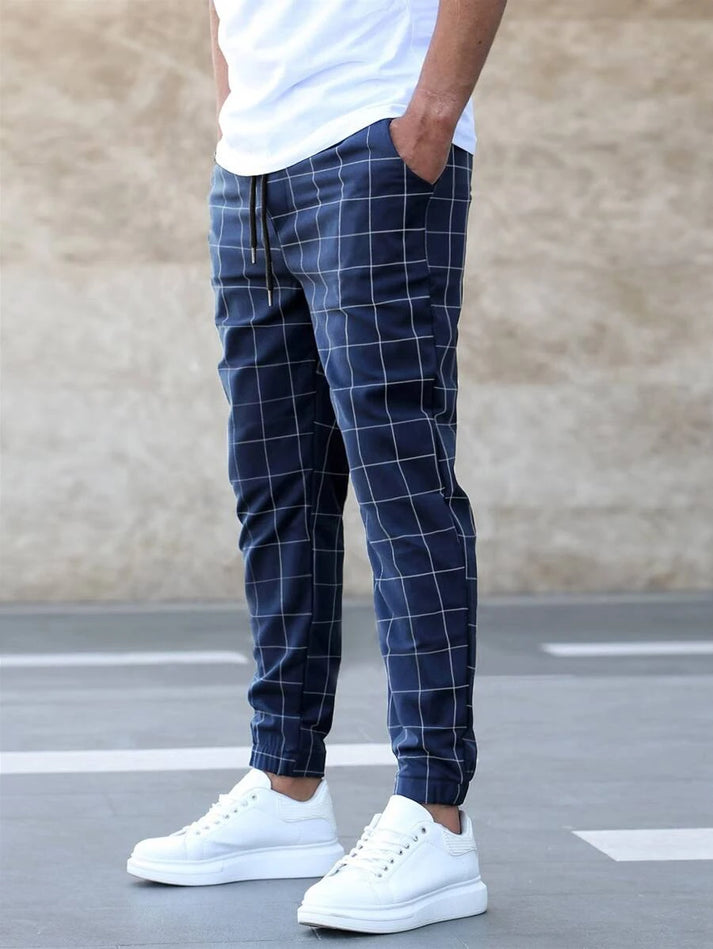 Mann in karierten Jogginghosen und weißen Sneakers vor beigem Hintergrund.