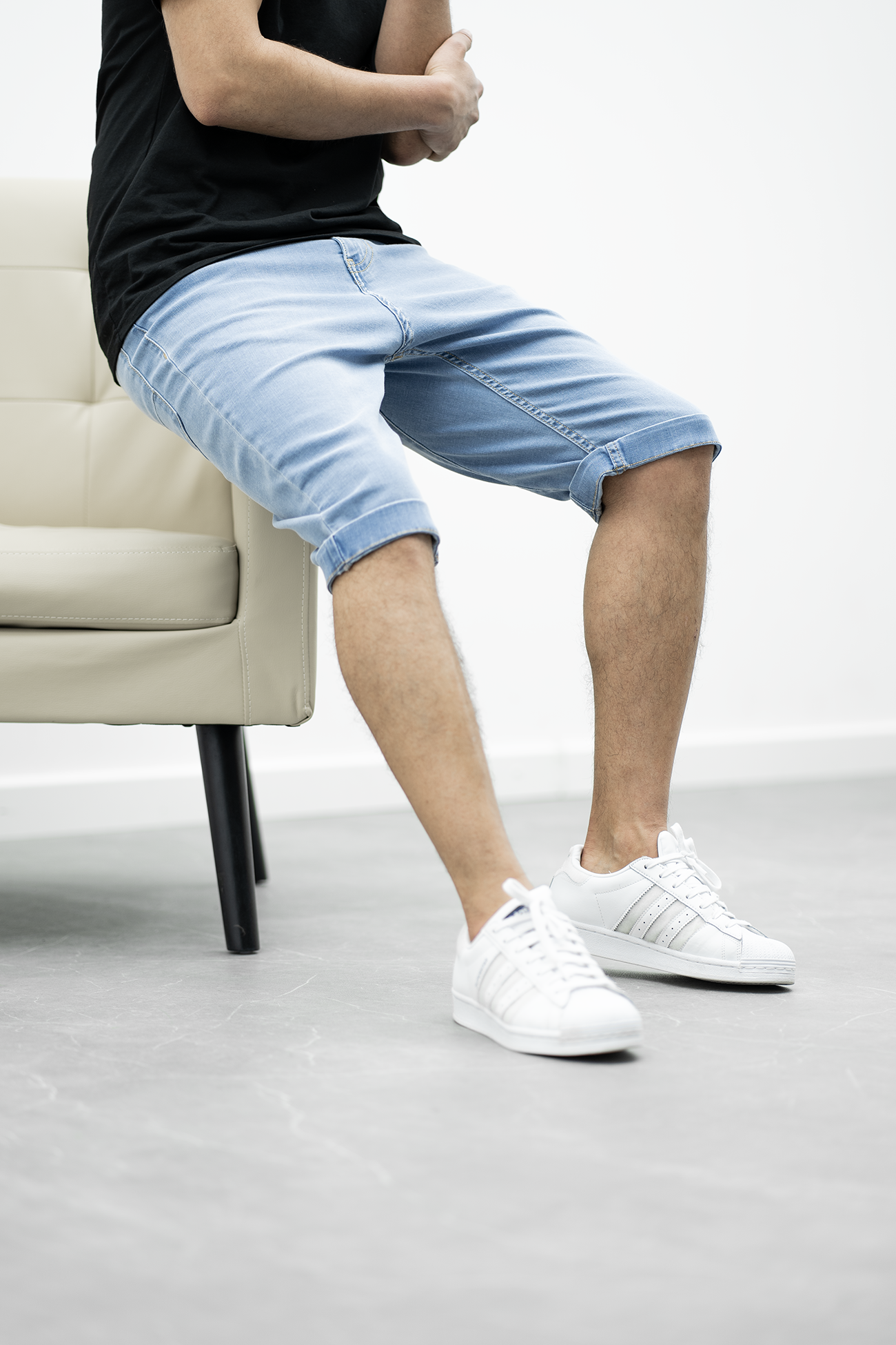 Mann in hellblauen Jeans-Bermudashorts, sitzt auf beigem Sessel, trägt weiße Sneakers.