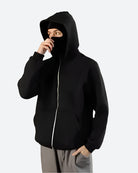 Person in schwarzem Hoodie mit Kapuze und Maske, grauer Hintergrund, modisch, urbaner Stil.