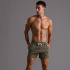Mann in gestreiften, olivgrünen Sportshorts, Fitnessmode, Performance-Activewear, grauer Hintergrund.