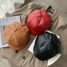 Drei modische Leder-Newsboy-Mützen in Rot, Braun und Schwarz auf einem Mantel. Hochwertige PU-Leder, ideal für Herbst und Winter.