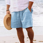 Mann in hellblauer Leinen-Shorts am Strand, hält Strohhut, Sommermode, Freizeitkleidung.