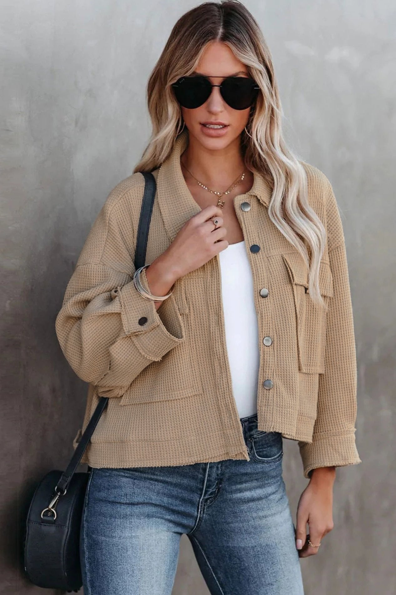 Frau in beiger Strickjacke, weißem Top und blauer Jeans, trägt schwarze Sonnenbrille und schwarze Tasche. Modetrends, Damenmode, lässiger Stil.