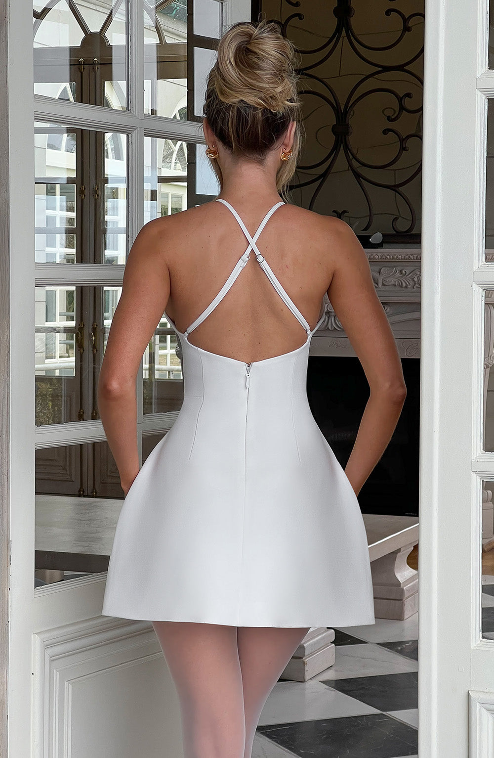 Frau in elegantem, weißem Cocktailkleid mit Kreuzträgern, steht vor einer Tür. Perfekt für Abendmode, stilvolle Partys, modische Eleganz.
