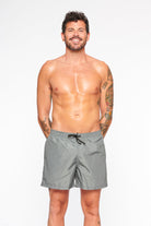 Mann in grauer Badehose, lächelnd, mit Tattoos auf den Armen. Sommermode, Herrenbadebekleidung, Strandoutfit, stilvolle Badeshorts.