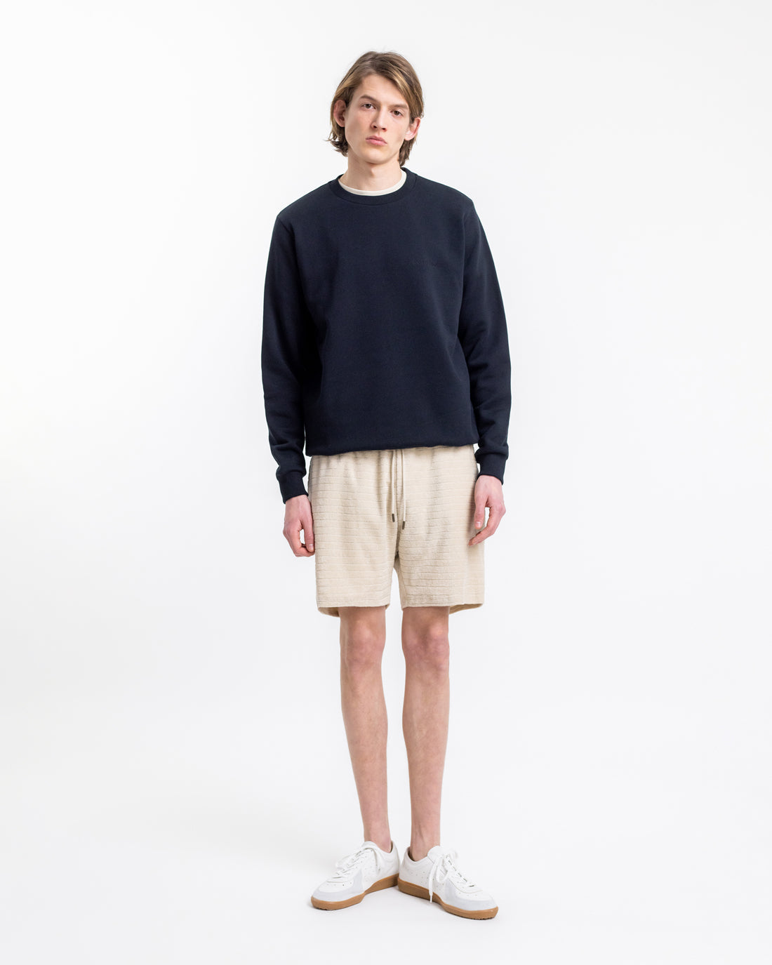 Junger Mann in lässigem Outfit: Dunkler Pullover, beige Shorts, weiße Sneaker. Minimalistischer Stil, moderne Herrenmode, Sommerlook.