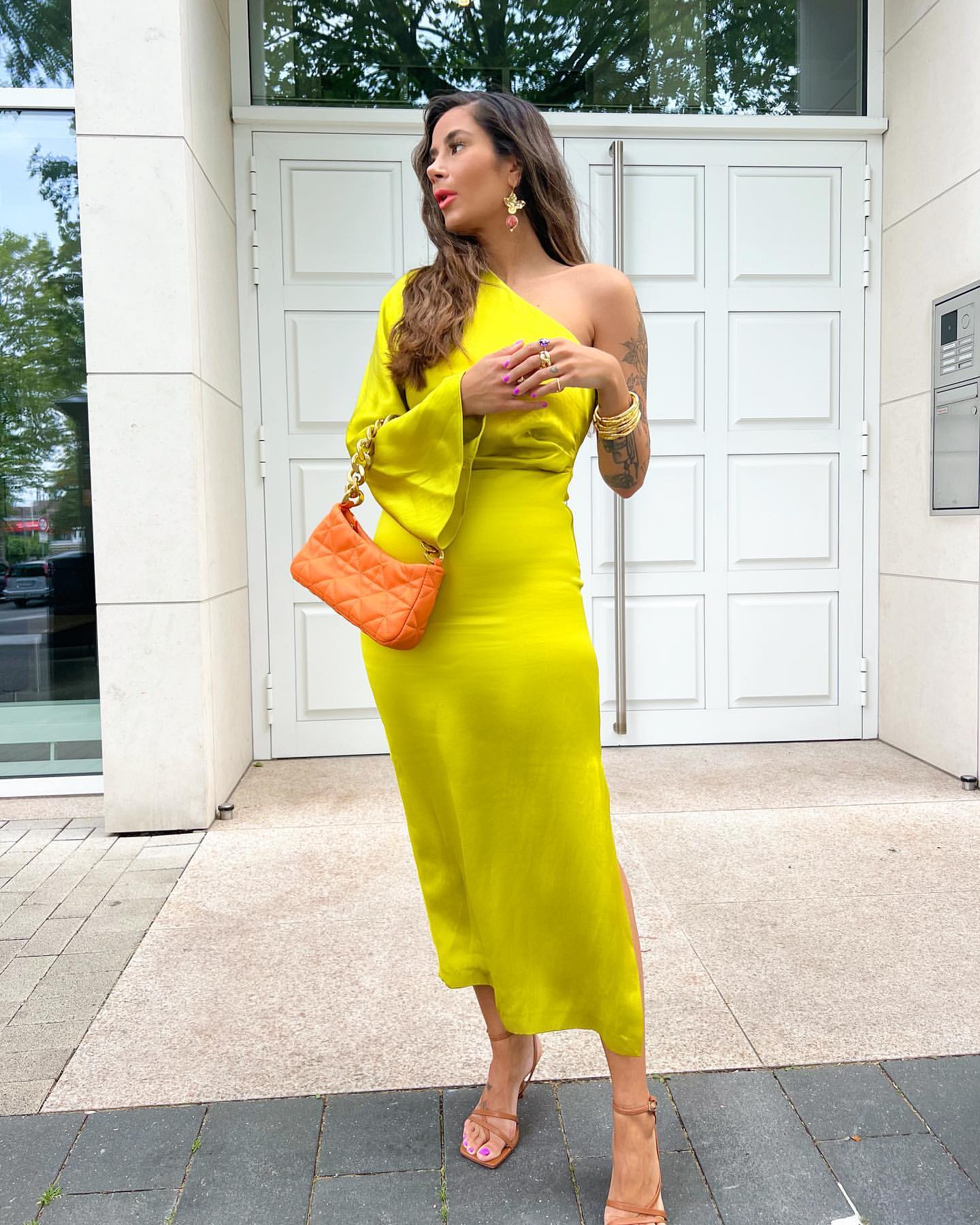 Frau in gelbem Kleid mit orangefarbener Tasche vor weißer Tür, modisch, elegant, Sommeroutfit.