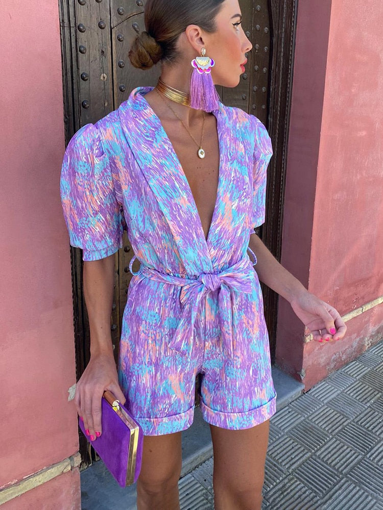 Frau in buntem, gemustertem Jumpsuit mit violetter Clutch vor rosa Wand. Modetrend, Sommeroutfit, stilvolle Accessoires, Streetstyle.