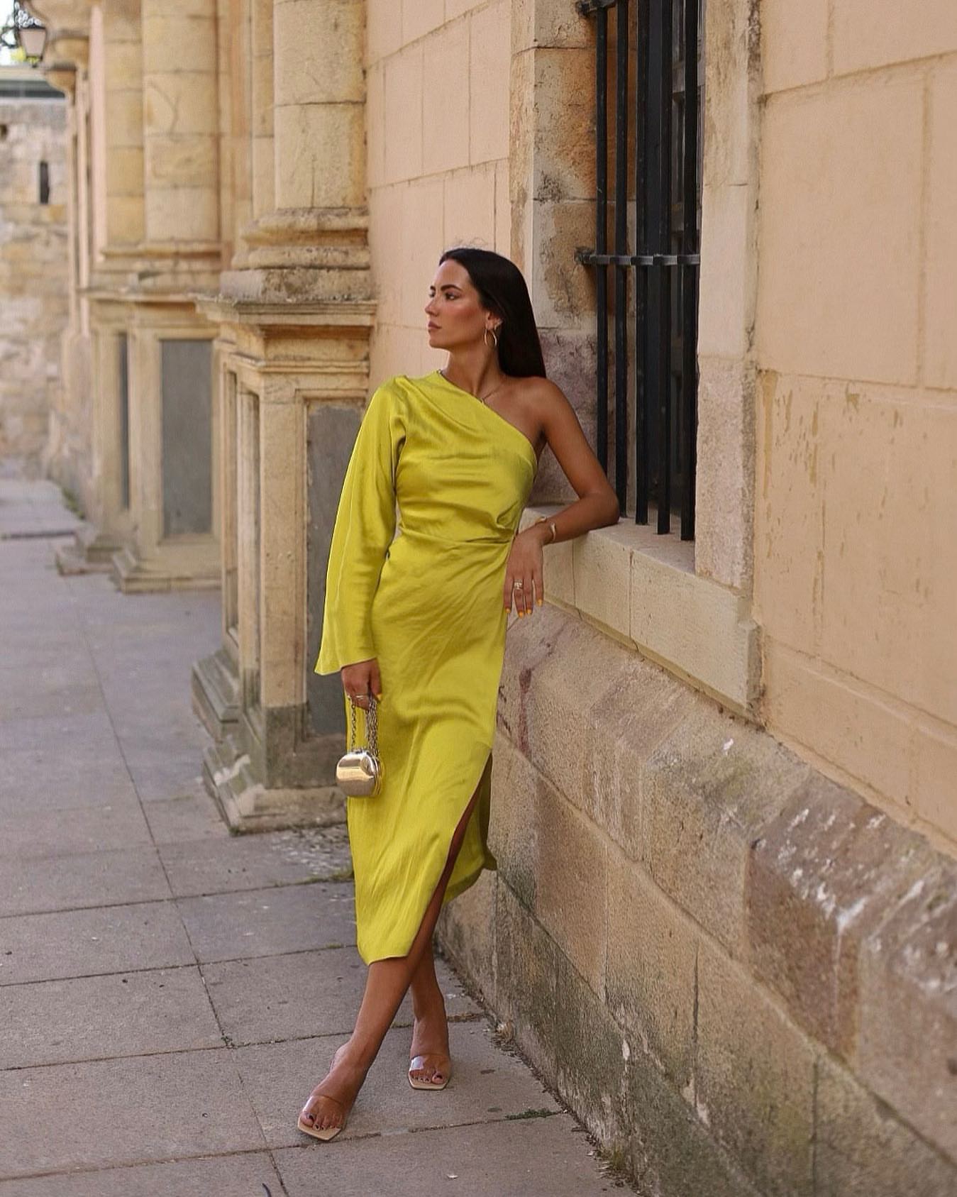 Frau in gelbem, einärmligem Kleid lehnt an Wand, hält goldene Handtasche, eleganter Look.