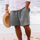 Mann am Strand in grauer Leinen-Shorts und weißem Hemd, hält Strohhut. Sommermode, Strandbekleidung, lässiger Stil, Herrenmode.