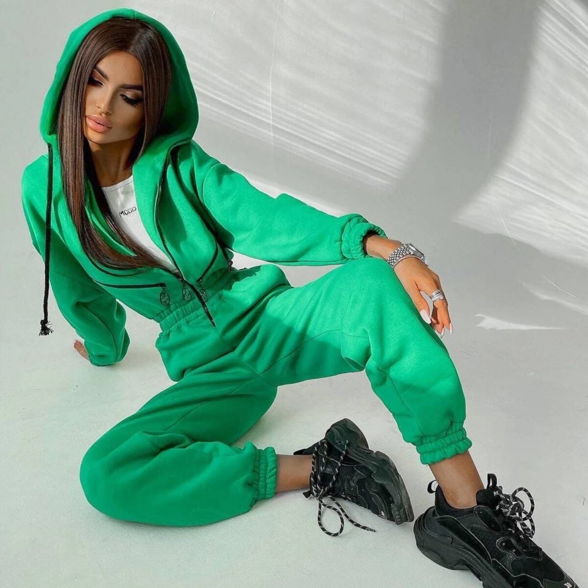 Frau in grünem Jogginganzug mit Kapuze, sitzt lässig, trägt schwarze Sneaker. Modetrend, Streetwear, bequeme Freizeitkleidung, stilvoll.