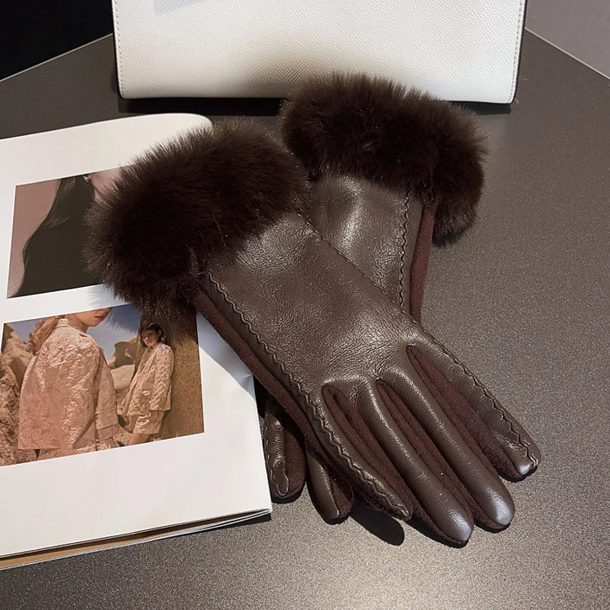 Damenlederhandschuhe mit Pelzbesatz auf Tisch, neben Modezeitschrift und weißer Tasche. Elegante Winteraccessoires, stilvoll und luxuriös.