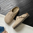 Beige Wildleder-Clogs mit Schnalle auf dunklem Holzboden. Komfortable, modische Schuhe für Damen und Herren. Ideal für Freizeit und Alltag.