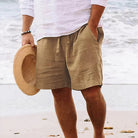 Mann am Strand in beigen Leinen-Shorts und weißem Hemd, hält Strohhut in der Hand.