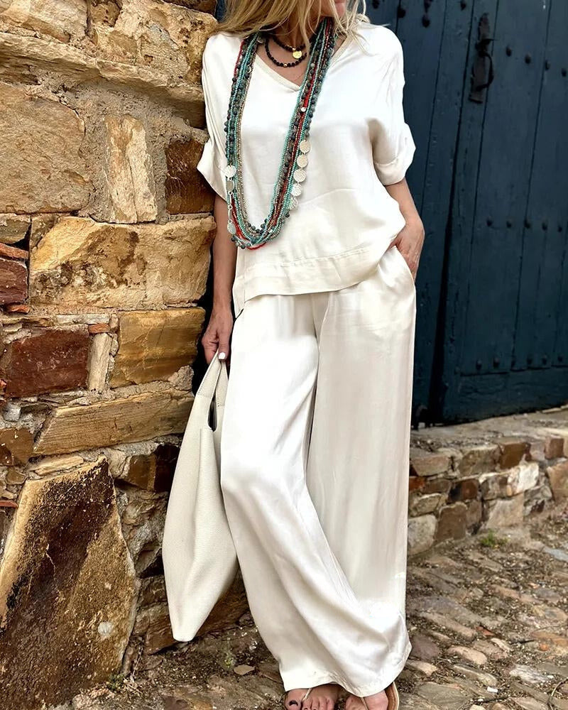Elegantes Damen-Outfit in Weiß mit lässiger Bluse und weiter Hose, ergänzt durch lange Halsketten. Perfekt für Sommermode und Boho-Stil.