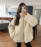 Frau in weißer Teddyjacke macht Selfie in modernem Wohnzimmer. Trendige Wintermode, gemütliche Jacke, stilvolle Inneneinrichtung, Smartphone.