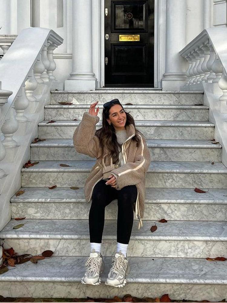 Frau in beiger Jacke und Sneakers sitzt auf Marmortreppe vor eleganter Haustür. Herbstblätter, Mode, Streetstyle, urbane Kulisse.
