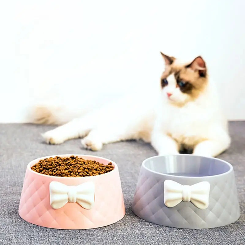 Rosa und graue Katzennäpfe mit Schleifen-Design, gefüllt mit Trockenfutter, vor einer flauschigen Katze. Perfekt für stilvolle Haustierfütterung.
