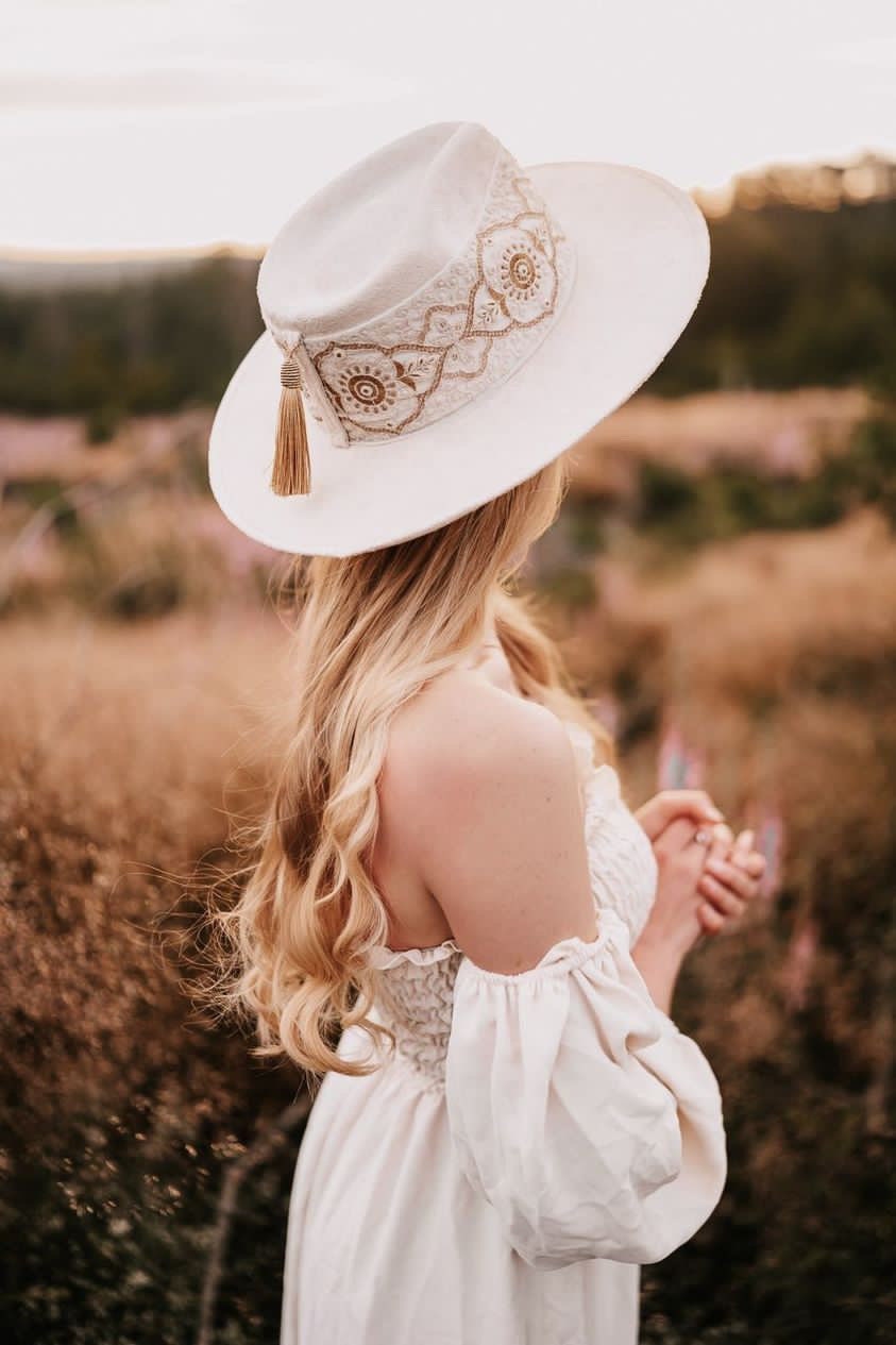 Frau mit weißem Spitzenkleid und besticktem Hut in blühender Wiese. Romantische Mode, Boho-Stil, Sommeroutfit, Naturkulisse.