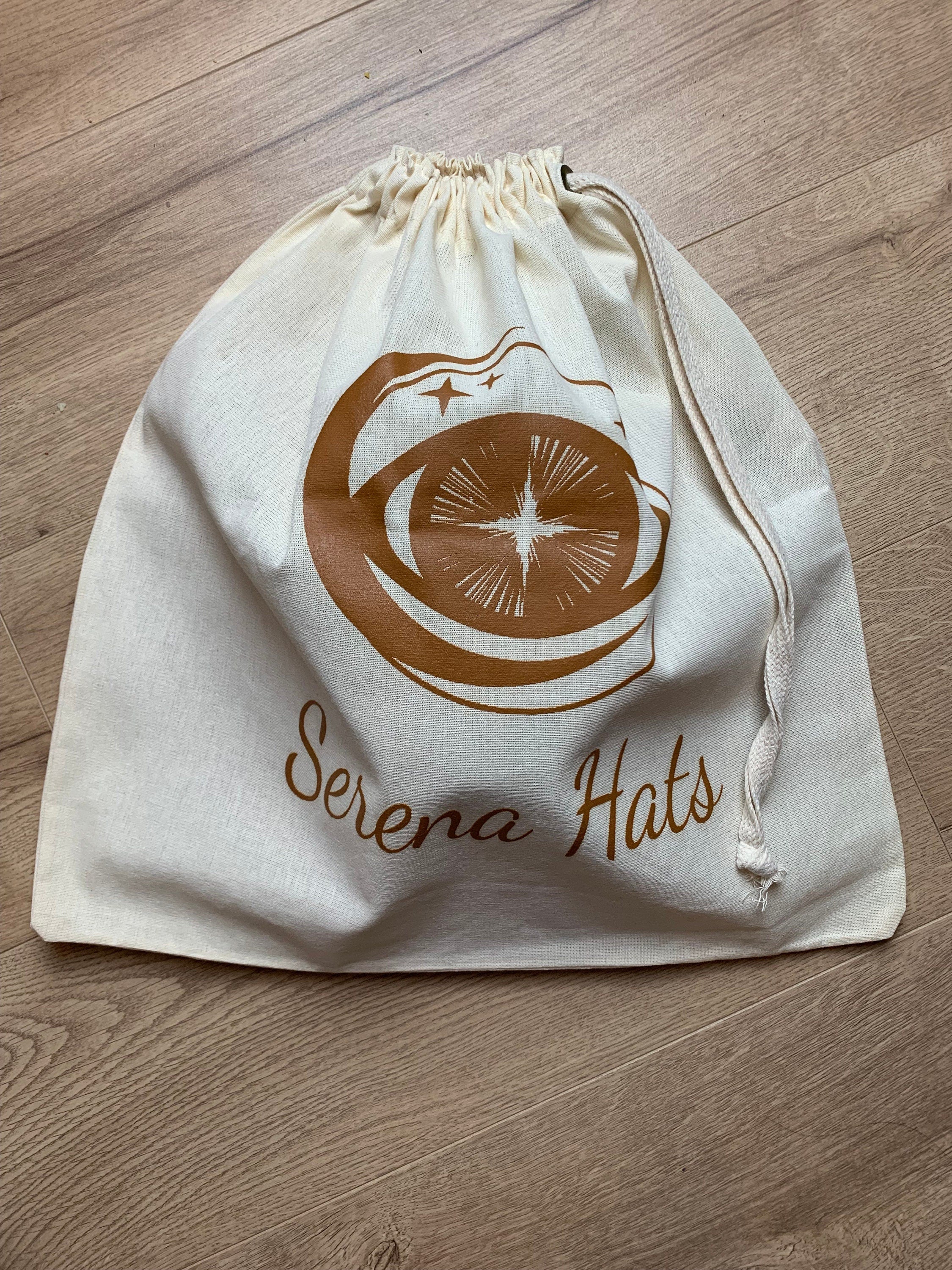 Baumwolltasche mit Kordelzug, bedruckt mit "Serena Hats" Logo in Braun. Perfekt für umweltfreundliche Aufbewahrung und Transport von Hüten.