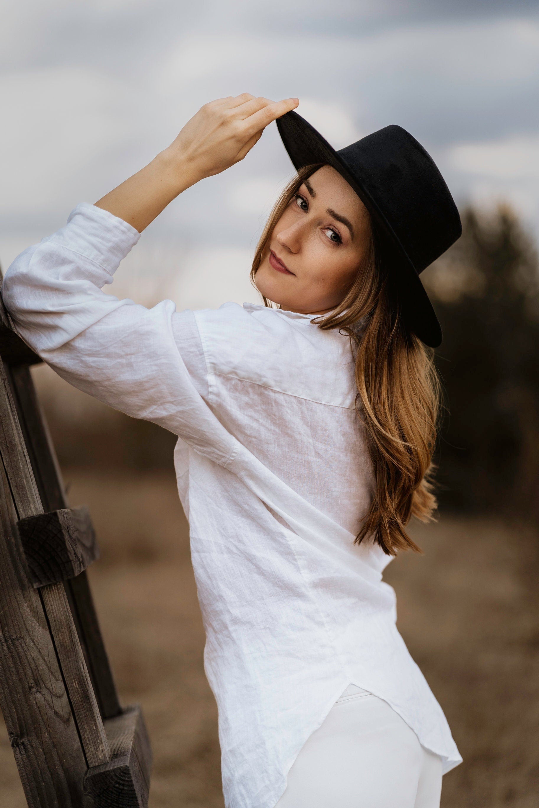 Frau in weißem Hemd und schwarzem Hut lehnt an Holzzaun, schaut über die Schulter. Mode, Outdoor, Herbst, lässiger Stil, Porträtfotografie.