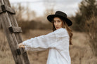Frau in weißem Leinenhemd und schwarzem Hut lehnt an Holzleiter im Freien. Mode, Natur, Herbst, stilvoll, lässig, Outdoor-Fotografie.