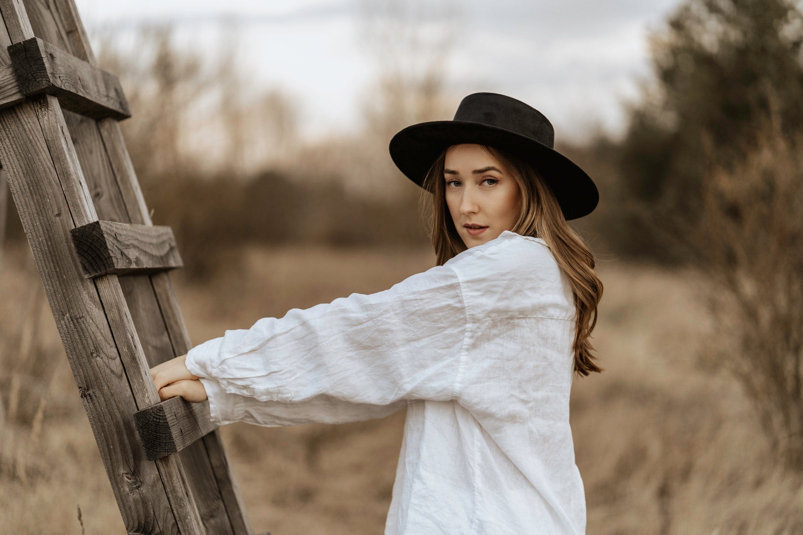 Frau in weißem Leinenhemd und schwarzem Hut lehnt an Holzleiter im Freien. Mode, Natur, Herbst, stilvoll, lässig, Outdoor-Fotografie.