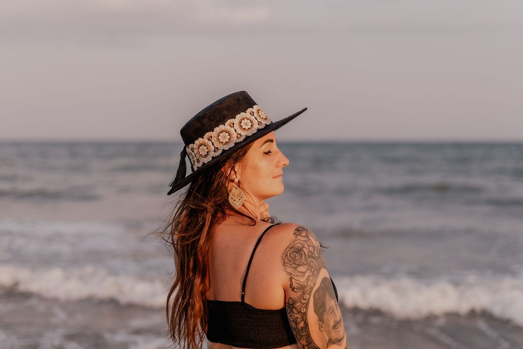 Frau am Strand mit schwarzem Hut und Blumenmuster, Tattoo auf dem Arm, Meer im Hintergrund. Sommermode, Strandaccessoires, Boho-Stil.