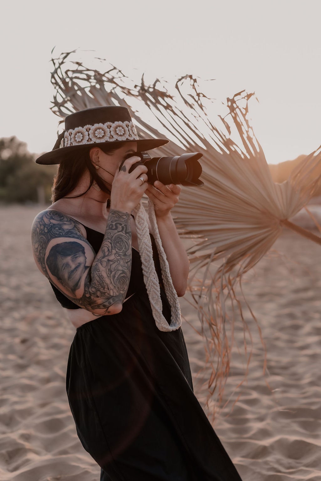 Frau mit Tattoo und Hut fotografiert am Strand bei Sonnenuntergang. Kamera, Fotografie, Strand, Sonnenuntergang, Tattoo, Hut, Natur.