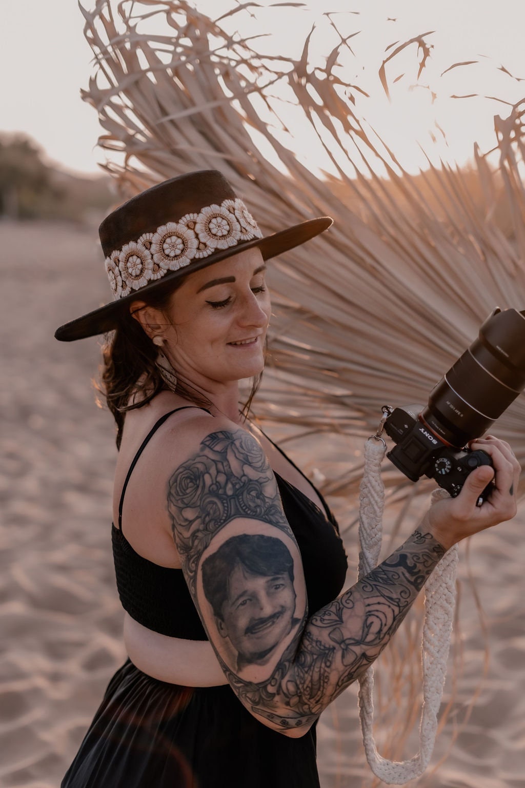 Frau mit Tattoo und Kamera am Strand, trägt einen Hut mit Blumenmuster. Perfekt für Fotografie, Strandmode und Tattoo-Designs.