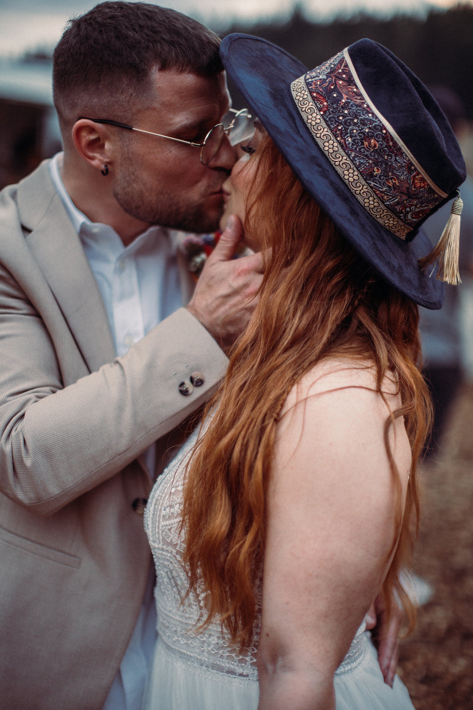 Paar in eleganter Hochzeitskleidung, Mann im Anzug küsst Frau mit langem Haar und Hut. Romantische Hochzeit, Boho-Stil, Outdoor-Setting.