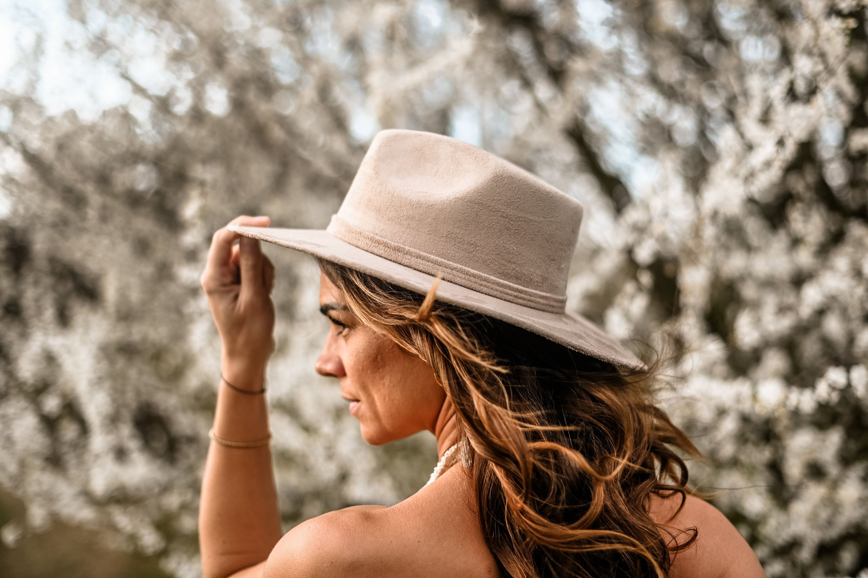 Frau mit beigem Hut vor blühendem Baum. Modischer Filzhut, Frühlingsmode, Outdoor-Fotografie, stilvolles Accessoire, Naturhintergrund.