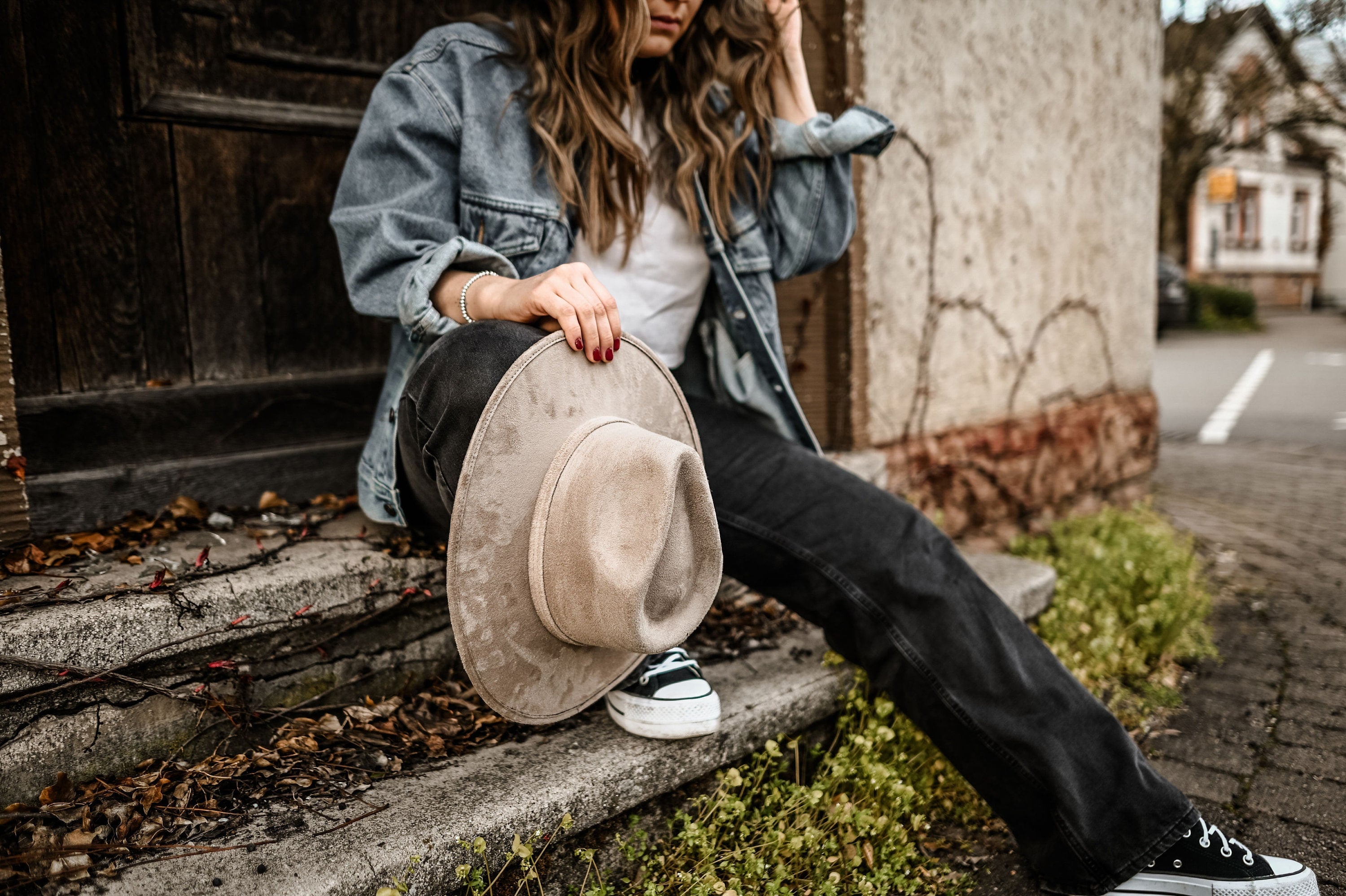 Frau in Jeansjacke und schwarzer Hose hält beigen Cowboyhut, sitzt auf Treppe. Lässiger Streetstyle, urbaner Look, Modefotografie.
