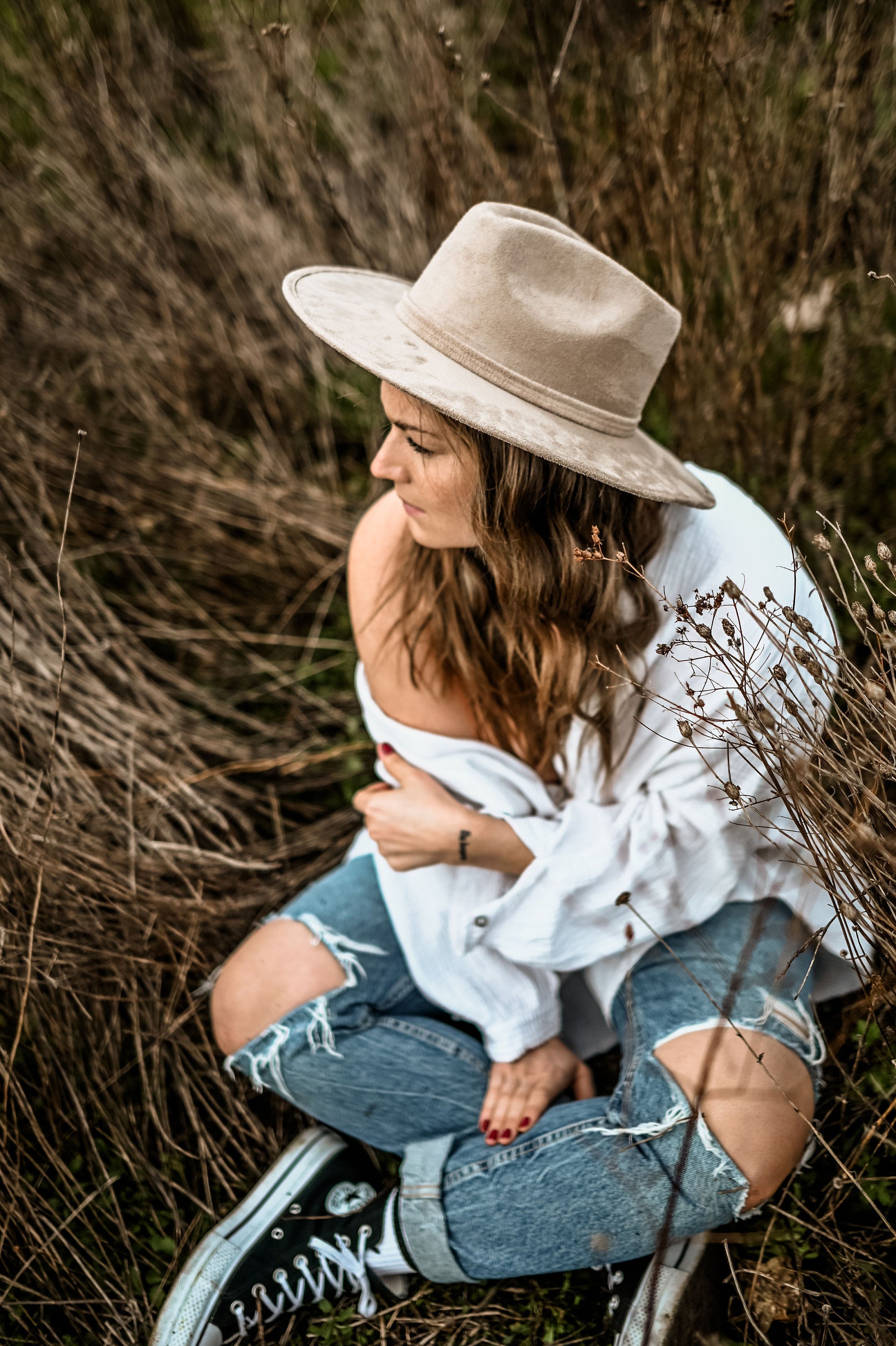 Frau in zerrissenen Jeans und weißem Hemd, sitzt im Gras, trägt beigen Hut. Lässiger Boho-Stil, Natur, Modefotografie, Outdoor-Porträt.