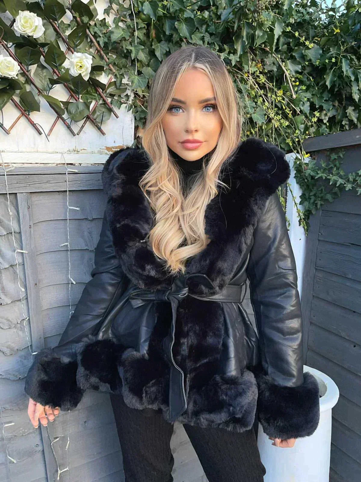 Frau in schwarzer Lederjacke mit Pelzkragen, blondes Haar, vor Holzwand mit Efeu und weißen Rosen. Modetrend, Wintermode, stilvoll.