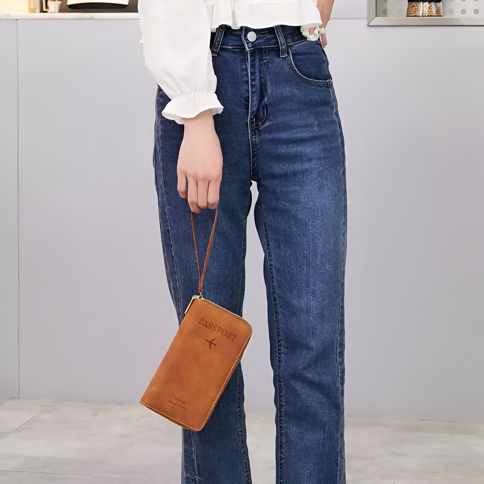 Frau mit blauer Jeans und weißer Bluse hält braune Leder-Reisepasshülle mit Handgelenkschlaufe.