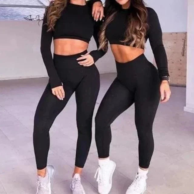Zwei Frauen in schwarzen Sportsets, bestehend aus Crop-Tops und Leggings, posieren in einem modernen Fitnessstudio. Ideal für Yoga und Fitness.
