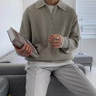 Mann in beigem Strickpullover hält ein englisches Wörterbuch. Stilvolle, moderne Herrenmode, gemütliches Wohnzimmer, minimalistische Einrichtung.